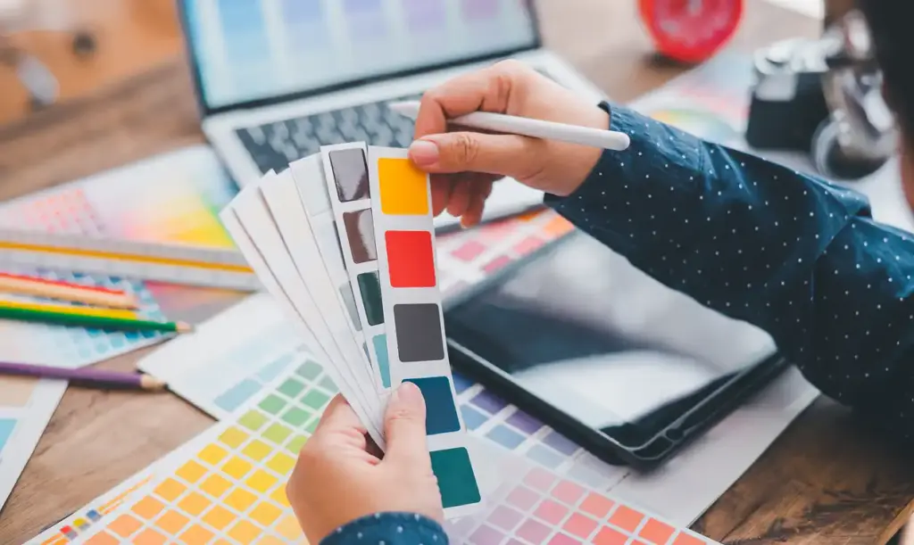 De handen van de ontwerper houden een kleurenpalet tegen een achtergrond van een tafel en computer.